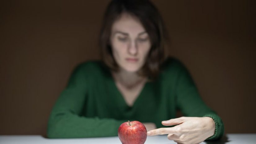 A woman in green thoughtfully observes a red apple on a white table indoors.